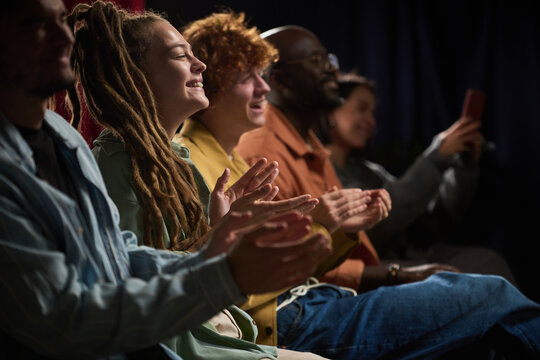 Group of diverse individuals of various ethnicities enjoying and applauding a performance, seated closely together in a darkened room sharing collective experience and connection