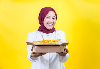 A cheerful asian woman in a hijab holds and showcasing a pizza box against a bright yellow backdrop