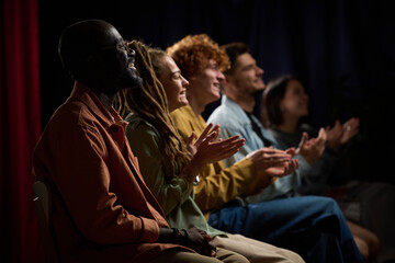 Group of diverse individuals applauding a performance with joyful expressions while seated in a dimly lit environment, highlighting togetherness and enjoyment