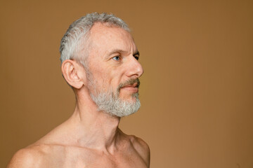 Obraz premium Side view of caucasian mature middle-aged shirtless man with grey hair and beard looking aside isolated in dark beige background copy empty space. Male beauty concept