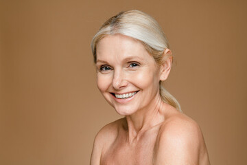 Headshot of gorgeous beautiful middle-aged woman with pure and healthy skin and blonde hair, stands isolated on brown beige background, mature lady looks at camera and smiles, copy space