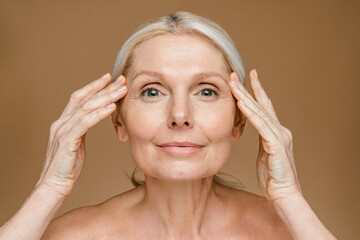 Portrait of beautiful mature blonde woman touching her face, posing over beige brown studio...