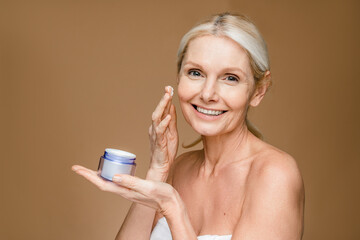Smiling tender woman holds jar of cream with anti aging effect applies on face has healthy glowing...