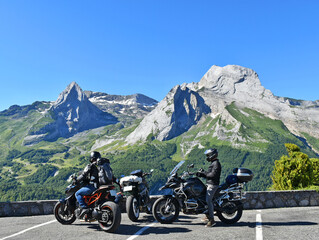 Motorcyclists in protective gear stop at a scenic mountain viewpoint with rugged peaks and green valleys