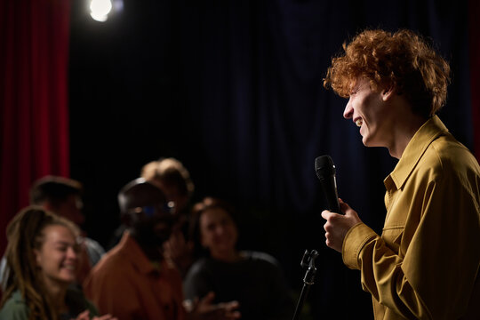 Person with curly hair holding microphone while performing stand-up comedy on stage. Audience consisting of diverse people enjoying show and laughing together in dark room