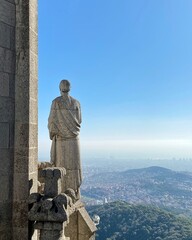 Fototapeta premium A statue stands on an outcrop in a temple, overlooking a panoramic view of a city below. The clear blue sky stretches out in the distance