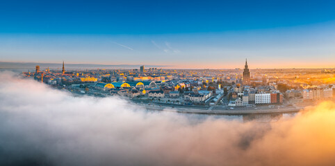 Fototapeta premium Thick fog covering Riga city in colorful sunrise colors. Drone view over old town, TV tower and river Daugava. Twilight colors in misty weather.