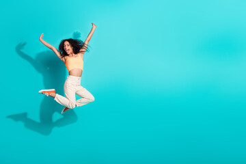 Fototapeta premium Excited young woman jumping joyfully on a vibrant turquoise background, expressing energy and happiness in casual summer fashion.