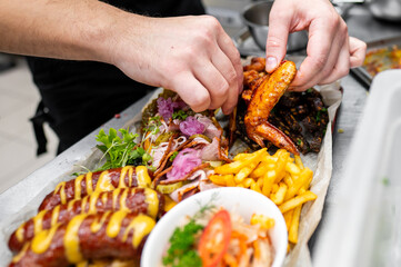 A close-up of hands preparing a delicious platter of assorted grilled meats, crispy fries, and fresh garnishes, showcasing vibrant colors and appetizing textures in a professional kitchen setting.