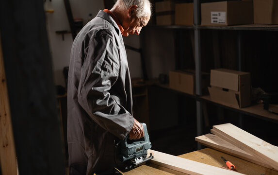 Senior retired carpenter sawing wooden plank using electric jigsaw. Carpenters work. Handyman woodworker in workshop. concept of skilled occupation and local artisans.