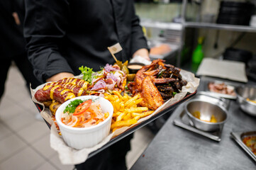 A chef holds a tray filled with a delicious assortment of grilled sausages, crispy fried potatoes, spicy chicken wings, fresh salad, and colorful garnishes in a bustling kitchen setting.