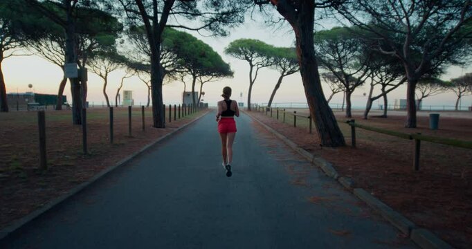 Parallax effect cinematic shot of young woman in pink running shorts and sports bra run in empty park on early morning, reach seaside promenade. Healthy habits, self care and sporty lifestyle