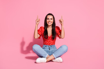Full size photo of pretty young woman sit floor direct fingers up empty space wear top isolated on pink color background