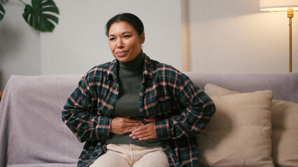 Upset Latin American woman bends massaging abdomen on sofa