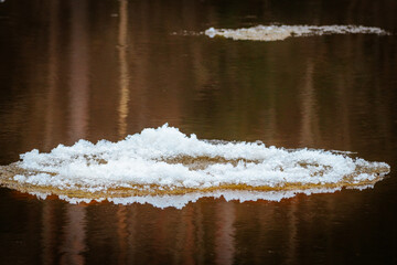 A floating piece of ice drifts on a calm river, reflecting in the dark water. The layered ice...