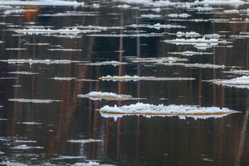 A floating piece of ice drifts on a calm river, reflecting in the dark water. The layered ice...