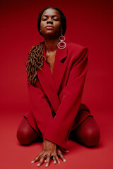 Portrait of poised African American woman kneeling on red floor against red backdrop wearing...