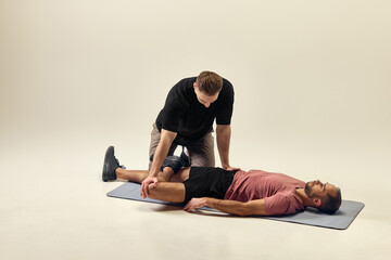 Trainer guiding man through spinal twist stretch, stabilizing leg while improving mobility against light studio background. Concept of rehabilitation, recovery, healthcare, physical therapy, sport