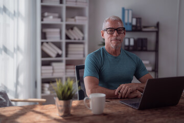 Confident mature businessman sitting at home office desk working remotely with laptop computer and drinking coffee, bookshelves and window in background