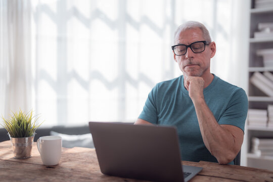 Focused mature man using laptop and thinking with hand on chin, working remotely from home sitting at wooden table with coffee cup and plant - Powered by Adobe