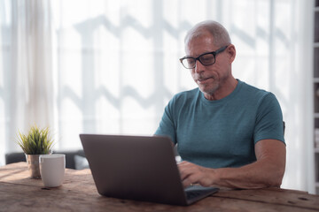 Fototapeta premium Focused mature man using laptop, typing on keyboard while working remotely from home office, enjoying comfortable and productive workspace with natural light