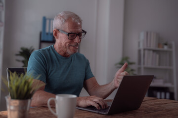 Fototapeta premium Happy senior man wearing glasses, working from home at his desk, engaged in a video call on his laptop, gesturing with his hand while smiling and enjoying the connection