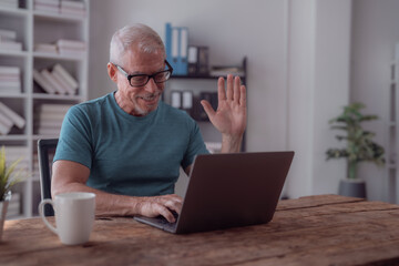 Smiling senior man wearing glasses waving at the camera while making a video call on his laptop from his home office, enjoying the convenience of working remotely
