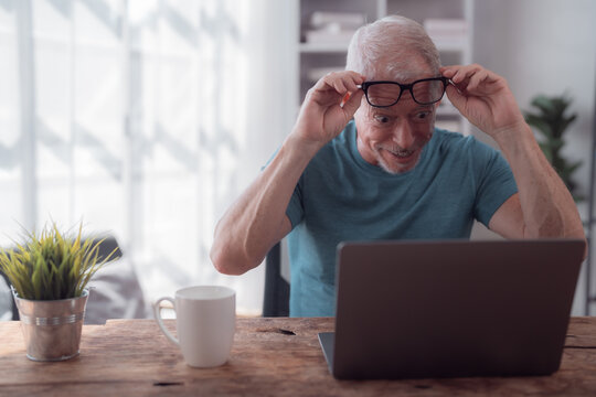 Surprised senior man adjusting glasses and staring at a laptop with a shocked expression, sitting at a wooden table with a cup of coffee, reacting to unexpected news