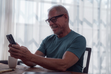 Happy retired man using smartphone for video call, browsing internet or reading messages sitting at wooden table at home with cup of coffee or tea
