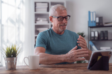 Cheerful mature man gesturing while video conferencing with colleagues or family on digital tablet, sitting at wooden table with cup of coffee in modern home office