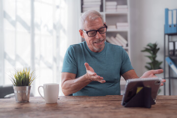 Happy retired man wearing eyeglasses having a video conference using a tablet at home, he is...