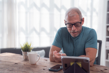 Smiling senior man wearing glasses gesturing while having a video call on a tablet at home, sitting at a wooden table with a smartphone and a coffee mug next to a small plant