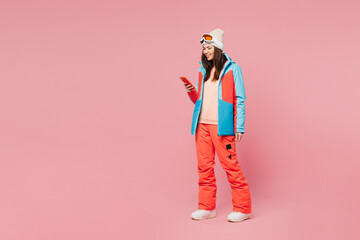 Full body skier young woman wear warm blue padded jacket ski goggles mask spend weekend winter season in mountains hold in hand use mobile cell phone isolated on plain pink background. Hobby concept. © ViDi Studio