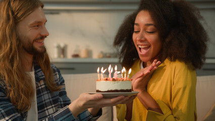 Happy couple multiracial multiethnic Caucasian man guy male boyfriend husband hold cake celebrate birthday African American woman girl female girlfriend wife blowing candle wishing at home couch hug