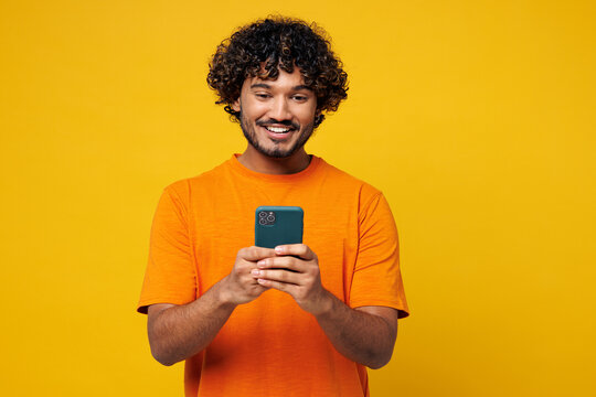 Young smiling happy student Indian man he wear orange t-shirt casual clothes hold in hand use mobile cell phone chatting online isolated on plain yellow background studio portrait. Lifestyle concept. - Powered by Adobe