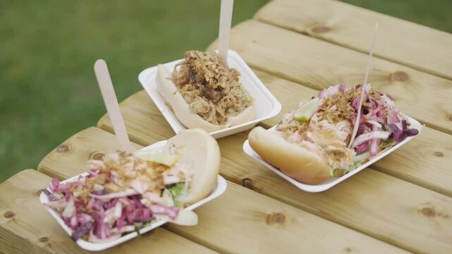 Three plates of food are on a wooden table. The food includes a salad, a hot dog, and a sandwich. The table is outdoors and the food is served on paper plates