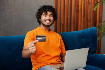 Young Indian man in orange casual clothes sits on blue sofa couch use laptop pc computer hold...