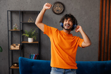 Young Indian man wears casual clothes sits on blue sofa couch listen music in headphones raise up...