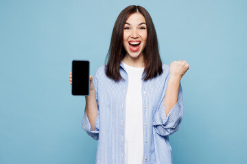 Young woman she wears shirt casual clothes hold in hand use mobile cell phone with blank screen workspace area do winner gesture isolated on plain pastel light blue cyan background. Lifestyle concept.