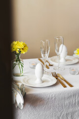 Happy Easter! Spring elegant table setting with white plates, egg cups, gold cutlery and vases with yellow flowers on a white tablecloth