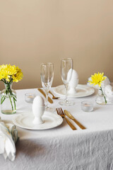 Happy Easter! Spring elegant table setting with white plates, egg cups, gold cutlery and vases with yellow flowers on a white tablecloth