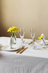 Happy Easter! Spring elegant table setting with white plates, egg cups, gold cutlery and vases with yellow flowers on a white tablecloth