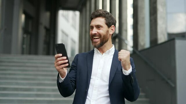 Happy excited businessman in a formal suit received great news on mobile phone standing on street near office building. Smiling shocked surprised entrepreneur reads positive good message on smartphone
