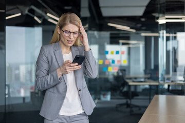 Bad news, negative online notification. Woman holding phone in hands disappointed and sad received message error using app. Businesswoman inside office at workplace nervous irritated.