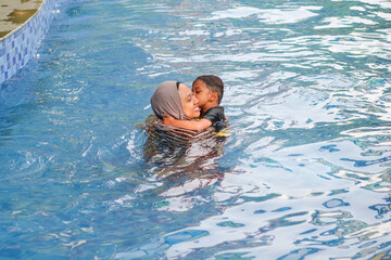 Beautiful young Asian muslim mother enjoying teaching her cute baby boy a first swimming lesson in a swimming pool