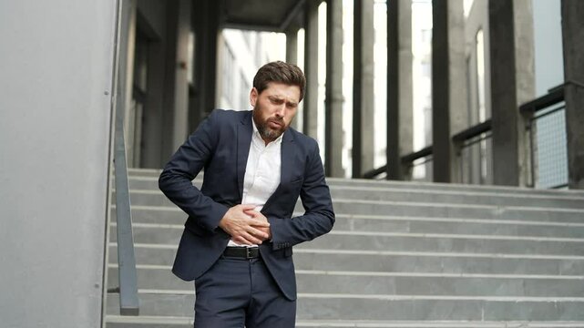 Upset businessman in formal suit feels stomach pain standing on street on the stairs of office building. Sick male has heartburn, gastritis or poisoning. Man suffers from overeating or constipation