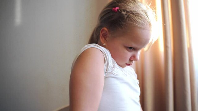 Young girl sits on chair, twists bare shoulder, looks down. Thoughtful expression, introspective moment, movement, physical awareness. Concept of child body language, emotions, concentration, motion