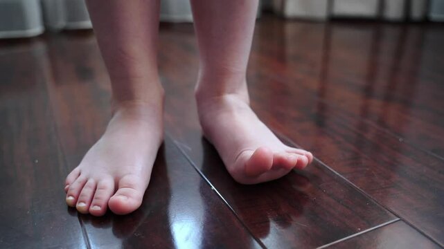 Girl stands barefoot on a wooden floor, shifting weight from foot to foot. Toes lift and wiggle, showing natural movement, flexibility. Smooth skin, arch, foot details visible in soft indoor lighting