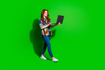 Smiling young woman with red hair using laptop over vibrant green background, wearing stylish...