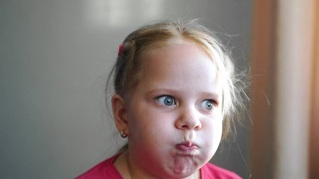 Close-up of young girl puffing out cheeks, pursing lips, moving mouth with funny expression. Cute artistic child making silly face. Blonde hair, blue eyes. Emotional, expressive, humorous moment